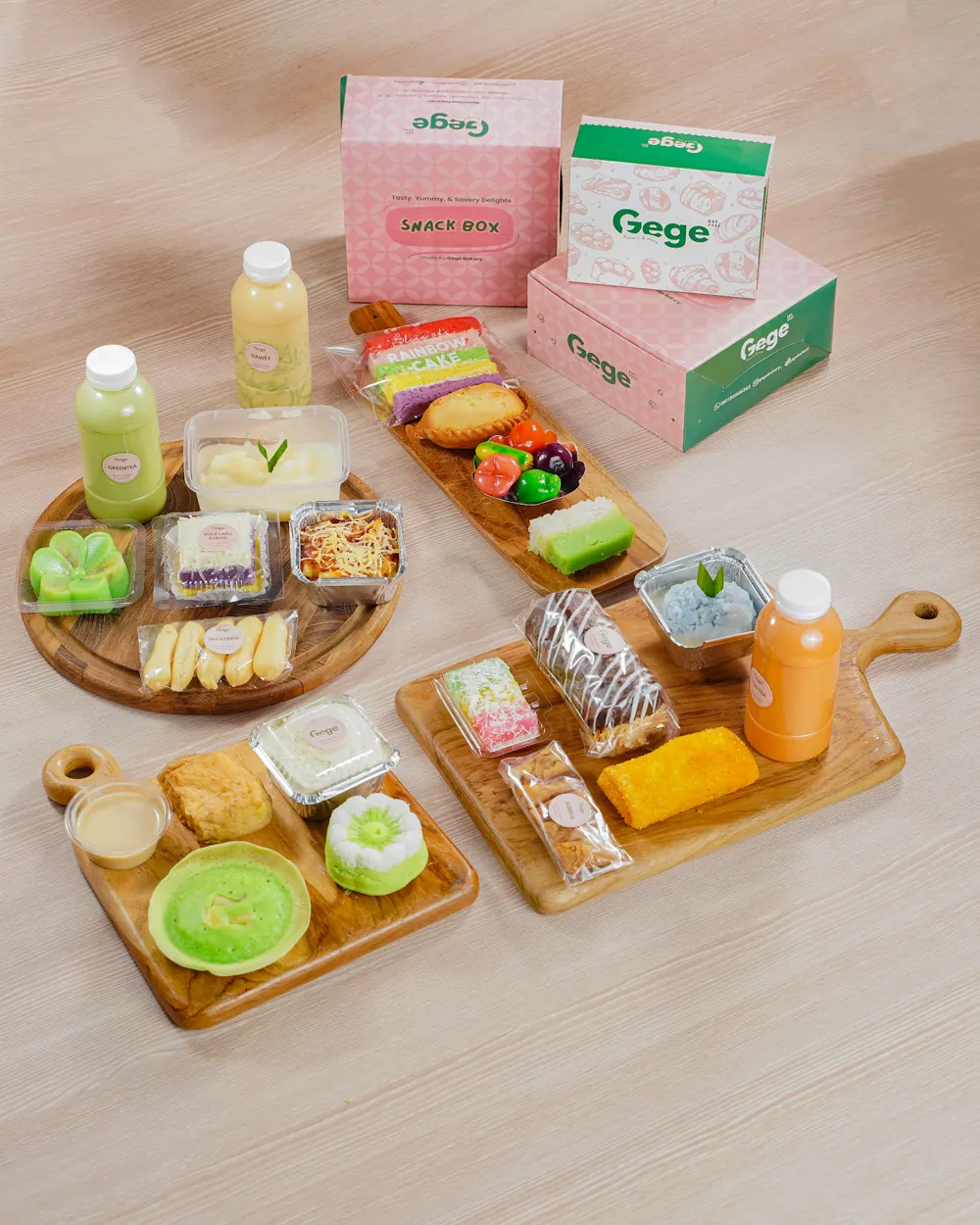 Aneka jajanan dan minuman di atas papan kayu, dengan kemasan snack box Gege berwarna merah muda dan hijau